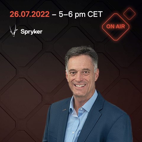 A man in a blue suit and light blue shirt smiles. Text above reads "26.07.2022 – 5-6 pm CET" with the "Spryker" logo and "ONAIR" on a dark geometric background.