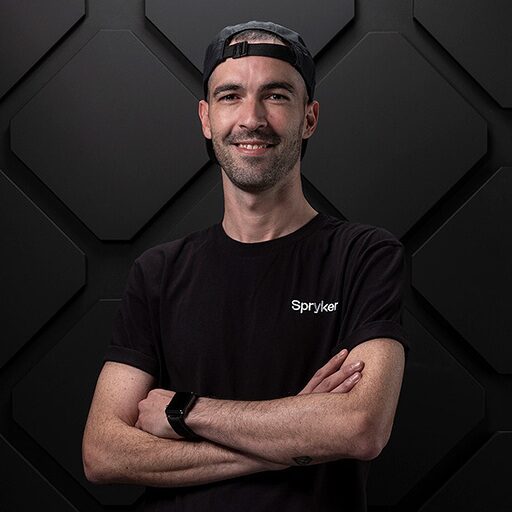 A man with a short beard and mustache smiles while standing with his arms crossed. He is wearing a black shirt featuring the "Spryker" logo and a black cap, embodying the future of AI enablement against a geometric dark background.