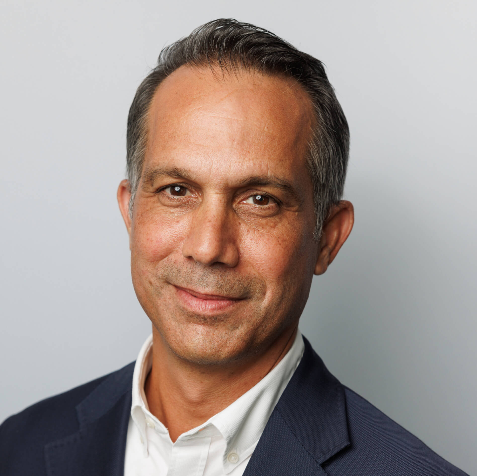 A man with short, grey hair wearing a navy blazer and white shirt smiles slightly against a plain background, seemingly ready for the automotiveIT Kongress.