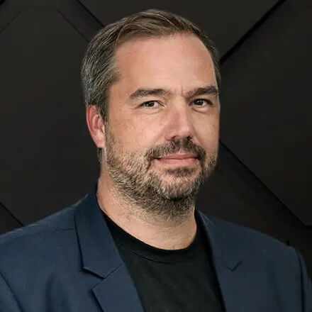 A man with short hair and a beard is wearing a black t-shirt and a dark blazer, facing the camera against a dark, textured background. His serious demeanor suggests he could be delivering an insightful keynote at the automotiveIT Kongress.