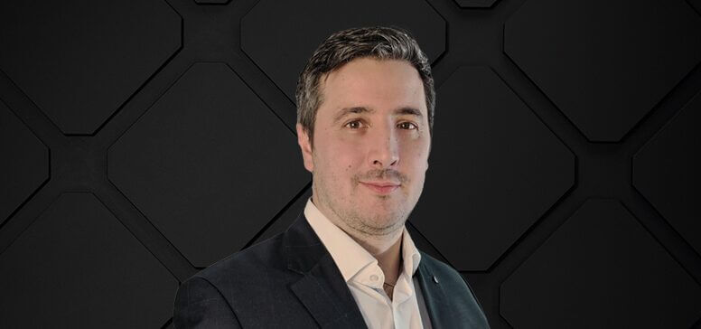 A man exuding leadership in a suit and light shirt stands confidently against a dark geometric background.