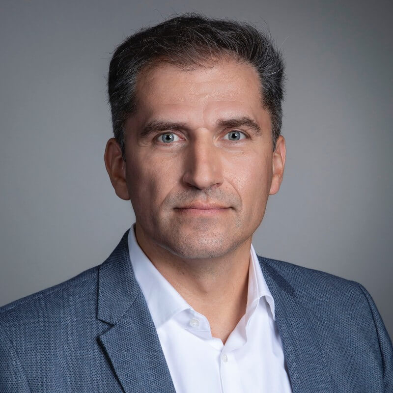 A middle-aged man with short dark hair wearing a blue blazer and white shirt, facing the camera with a neutral expression against a plain gray background, suitable for an OMR 2025 professional profile.