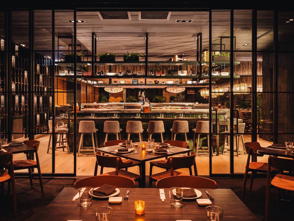 Dimly lit modern restaurant with wooden tables set for dining at Spryker's Sushi & Sake Social 2025. A bar with high stools is in the background, surrounded by shelves of plants and bottles, offering an innovative blend of B2B E-commerce and culinary excellence.