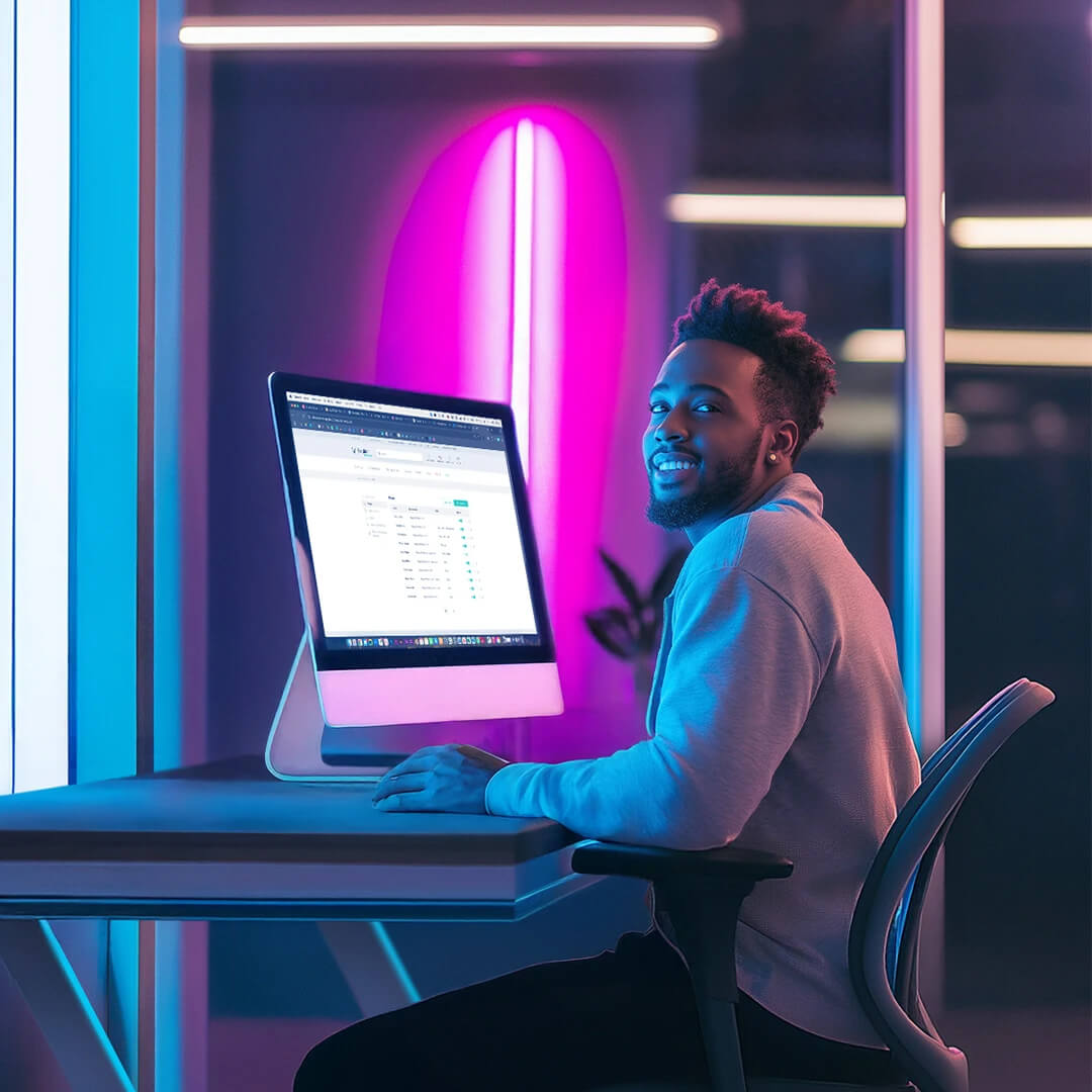 Person sitting at a desk, smiling, in front of a large computer monitor displaying spreadsheets. The room is illuminated with blue and purple lights.