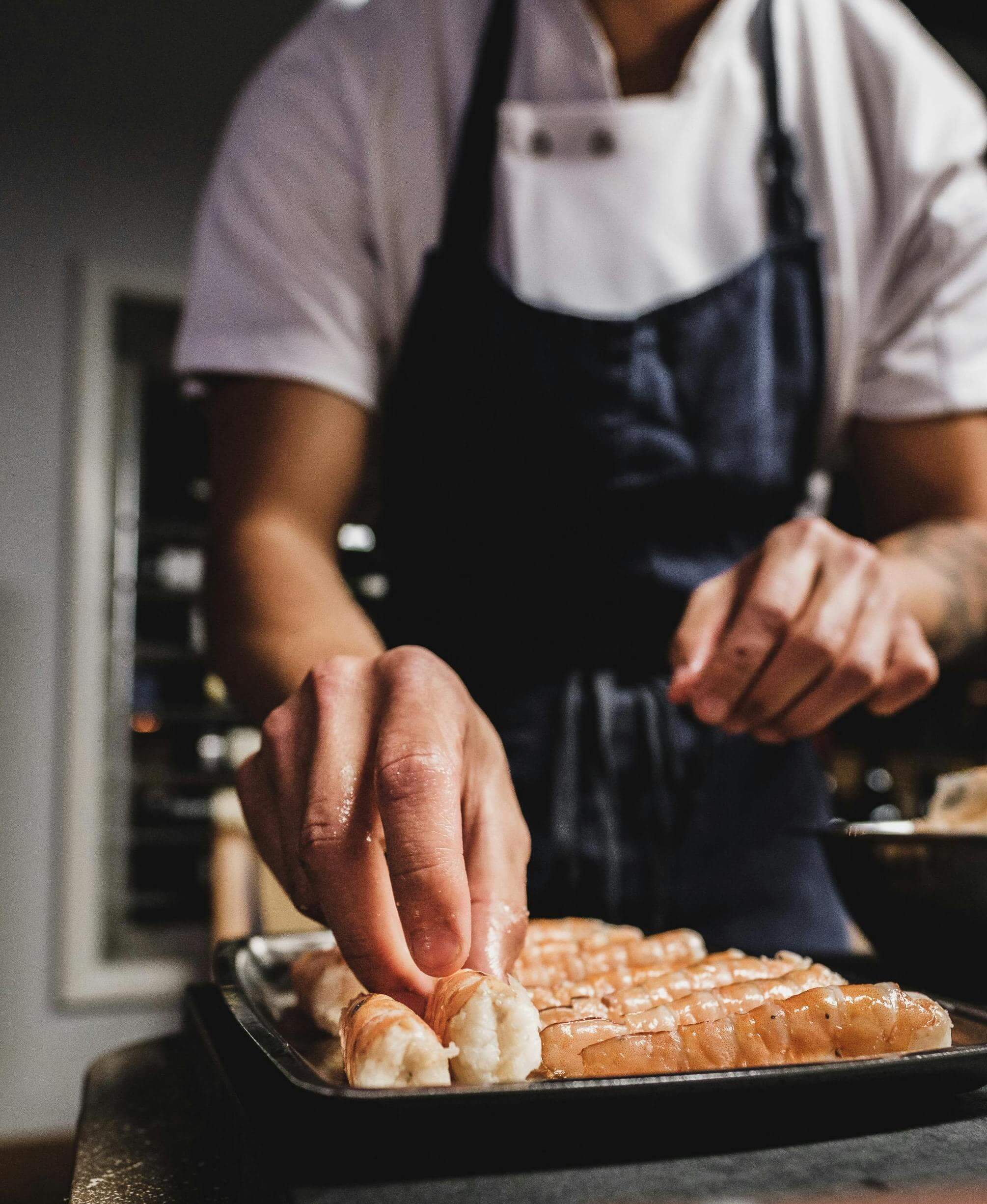 At Spryker's Sushi & Sake Social 2025, a chef in a white shirt and dark apron deftly prepares sushi rolls on a tray, showcasing culinary artistry that promises an unforgettable dining experience.