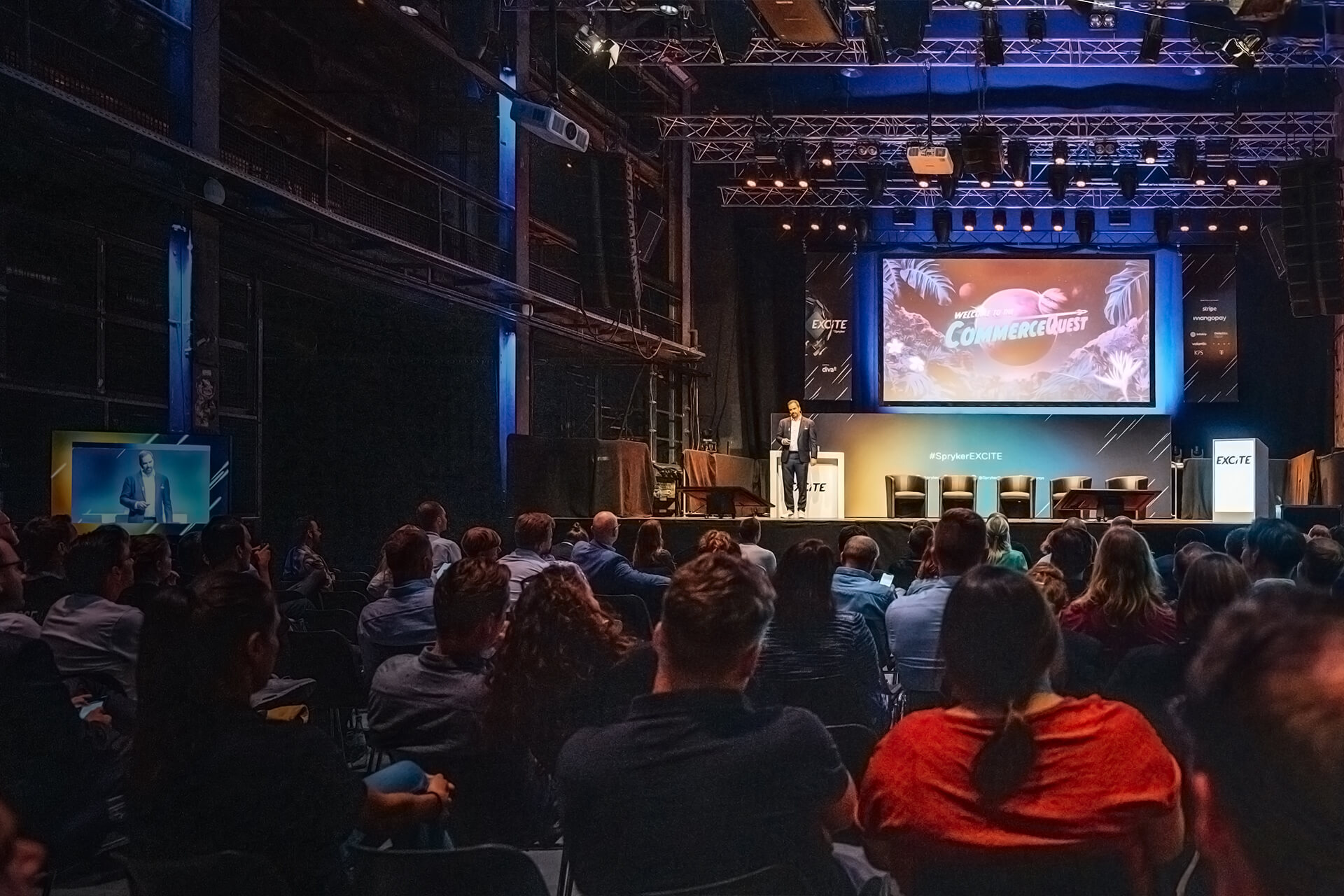 A speaker excites the audience on stage in a dimly lit auditorium, with a large screen behind displaying the word "COMMERCEGUEST" and hints of what’s to come in 2025.