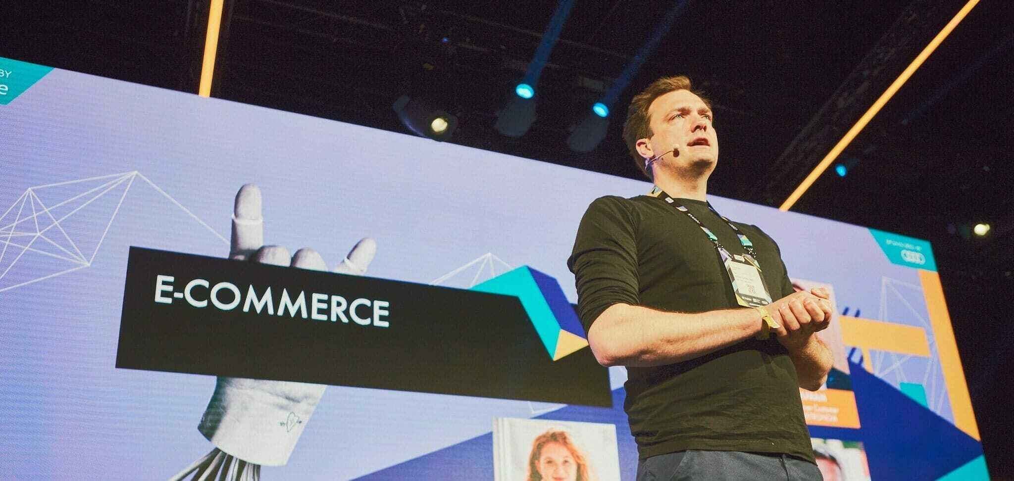 A speaker stands on a stage in front of a large screen displaying the word "E-COMMERCE" and graphics, as part of the OMR Festival 2022.