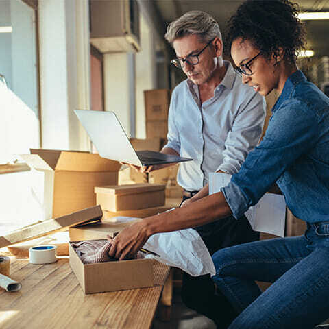 Two people are packing items into boxes on a table. The person on the left, likely coordinating through a b2b marketplace on their laptop, while the one on the right places an item into a box. There are packing supplies scattered across the table.