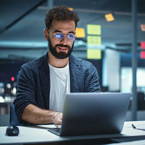 A man with glasses and a beard is sitting at a desk in an office, working on a laptop with composable commerce tools. There are sticky notes on the glass wall behind him.