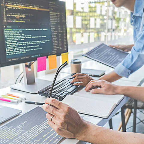 Two people collaborate at a desk with multiple computer monitors displaying code. One person holds a notebook, and the other holds a stylus, engaging in composable commerce solutions. The desk is adorned with sticky notes and stationery, indicating an active workspace.