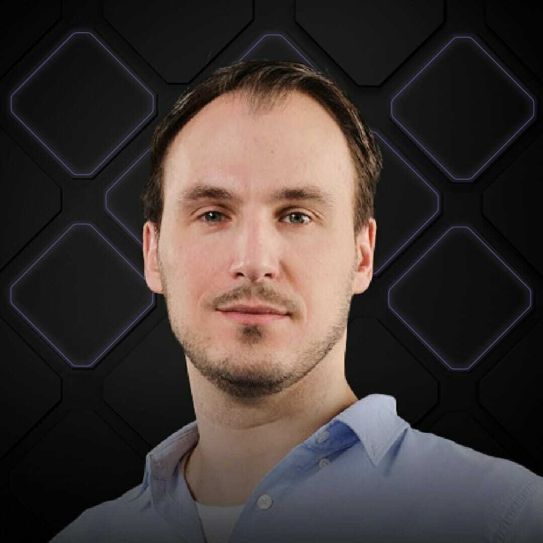 A man with short dark hair and a trimmed beard wears a light blue shirt at Spryker EXCITE 2022. The background consists of a black pattern with illuminated hexagonal shapes.