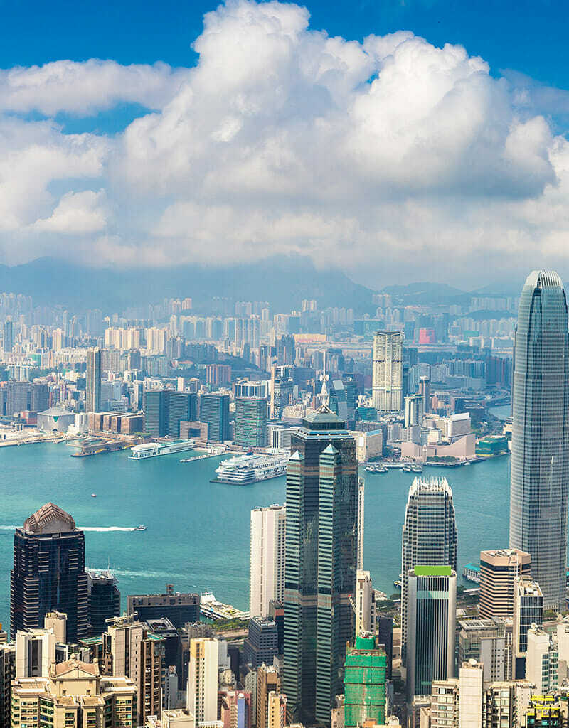 Panoramic view of a dense urban skyline with numerous high-rise buildings and skyscrapers near a large body of water, boats visible on the water, under a partly cloudy sky. In this bustling e-commerce hub in China, the city thrives day and night.