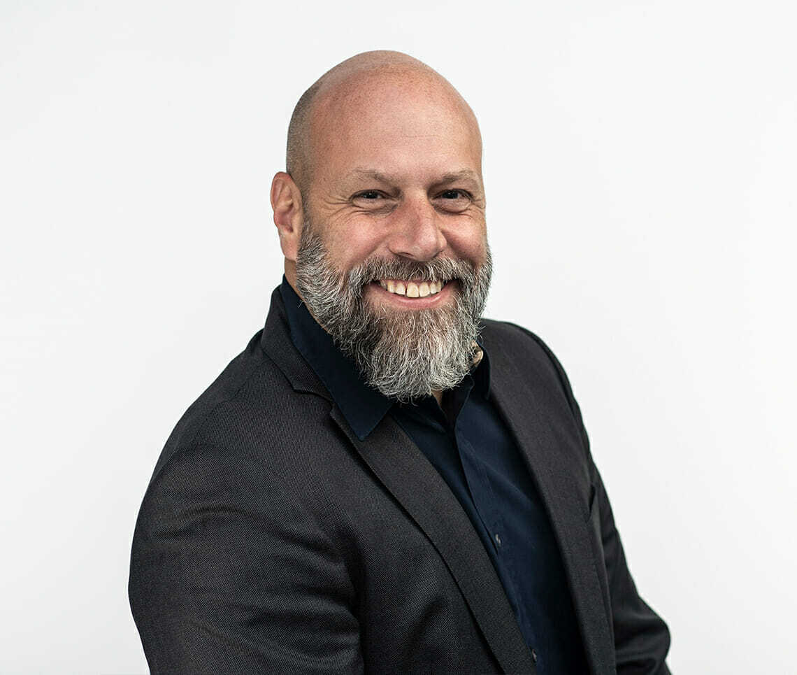 A bald man with a beard, smiling, wearing a dark blazer and shirt, poses confidently against a plain white background, embodying the professional spirit of Spryker at Shoptalk Europe.