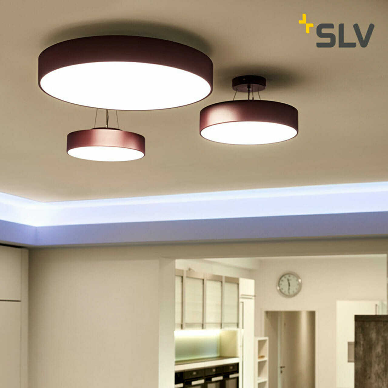 Three round ceiling lights with purple bases illuminate a modern, white kitchen featuring a clock on the wall. The lights are of different sizes, adding a touch of elegance to the space. The "SLV" logo in the upper right corner emphasizes their collaboration with a leading digital company.