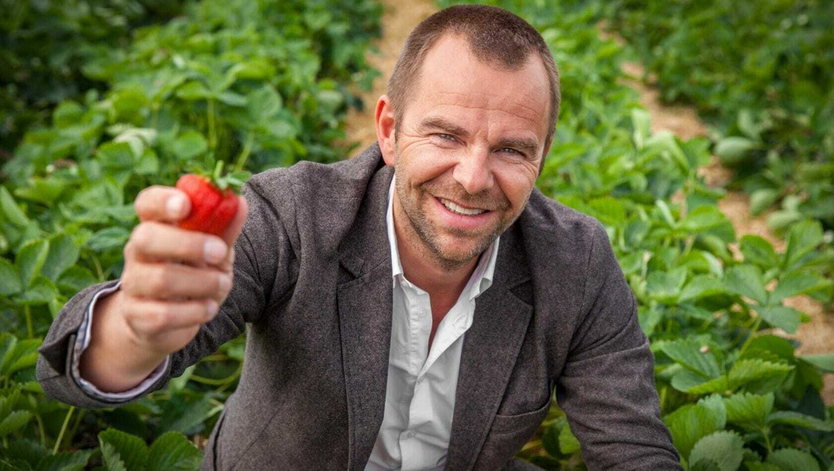 A man in a gray jacket and white shirt smiles while holding a strawberry in a field of greenery during the OMR Festival 2022.
