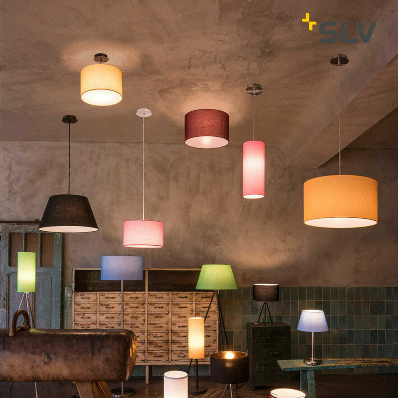 A variety of pendant and table lamps with different colored shades are displayed in a dimly lit room. The SLV logo is visible in the top right corner, subtly emphasizing the brand's innovative approach as a leading digital company.